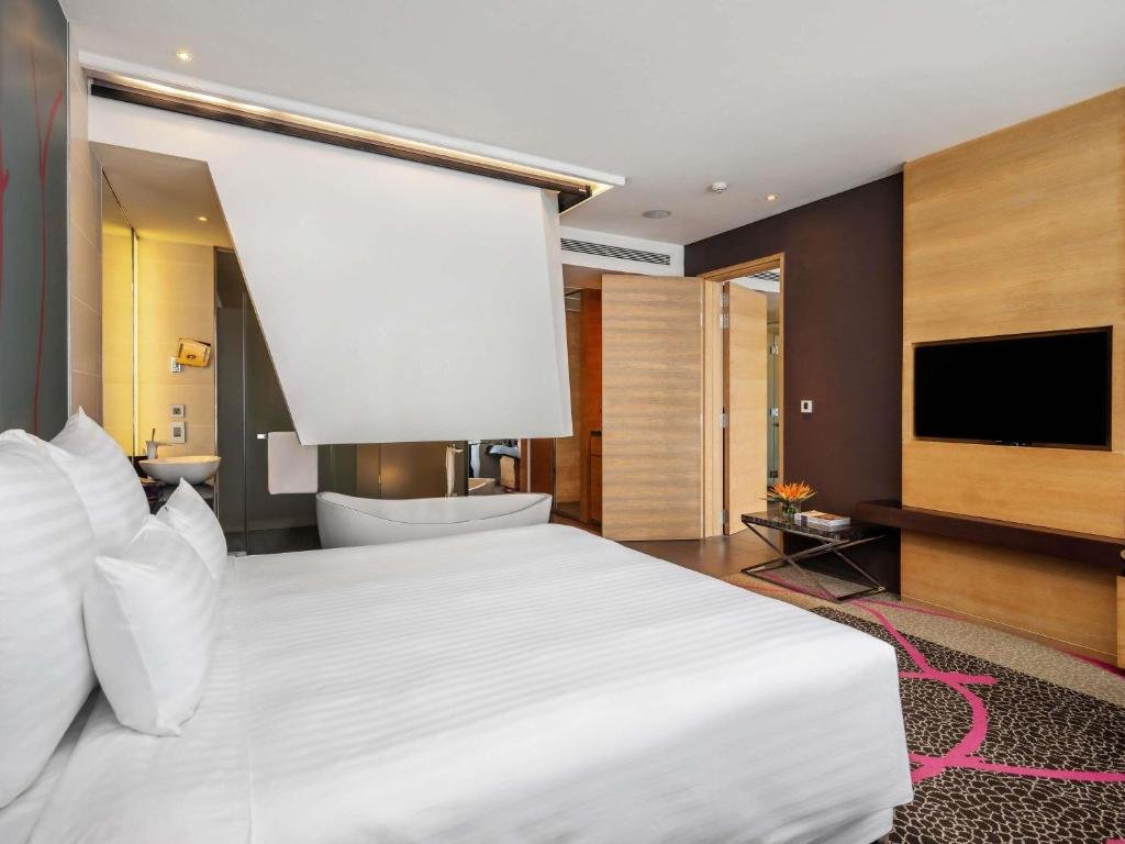 The king bed in the foreground, the freestanding bathtub and basin behind a partial wall, a wood-panelled TV wall, and a small side table with a bird-of-paradise flower
