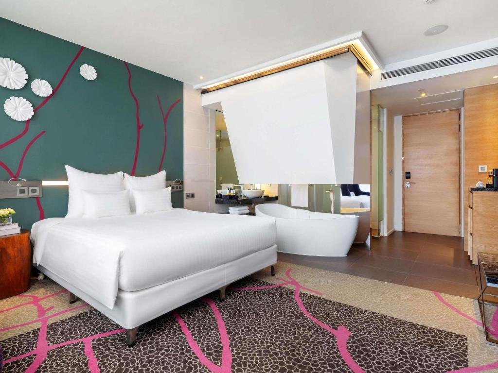 The room from beside the bed: a white freestanding bathtub under an angled white canopy, open to the bedroom, with a bowl basin and glass shower behind and the entry door on the right