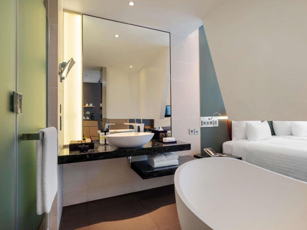 A white bowl basin on a black marble counter under a large mirror, the curved rim of the freestanding bathtub in front and the bed to the right