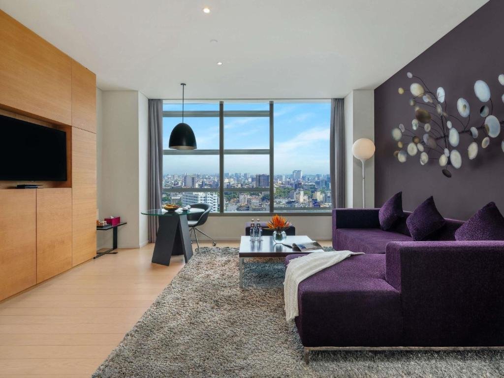 The Executive Suite living room at Pullman Saigon Centre: a purple sectional sofa on a shaggy grey rug, a metal disc sculpture on a plum wall, a TV set into oak panelling, and a round glass desk at a full-height window over the city