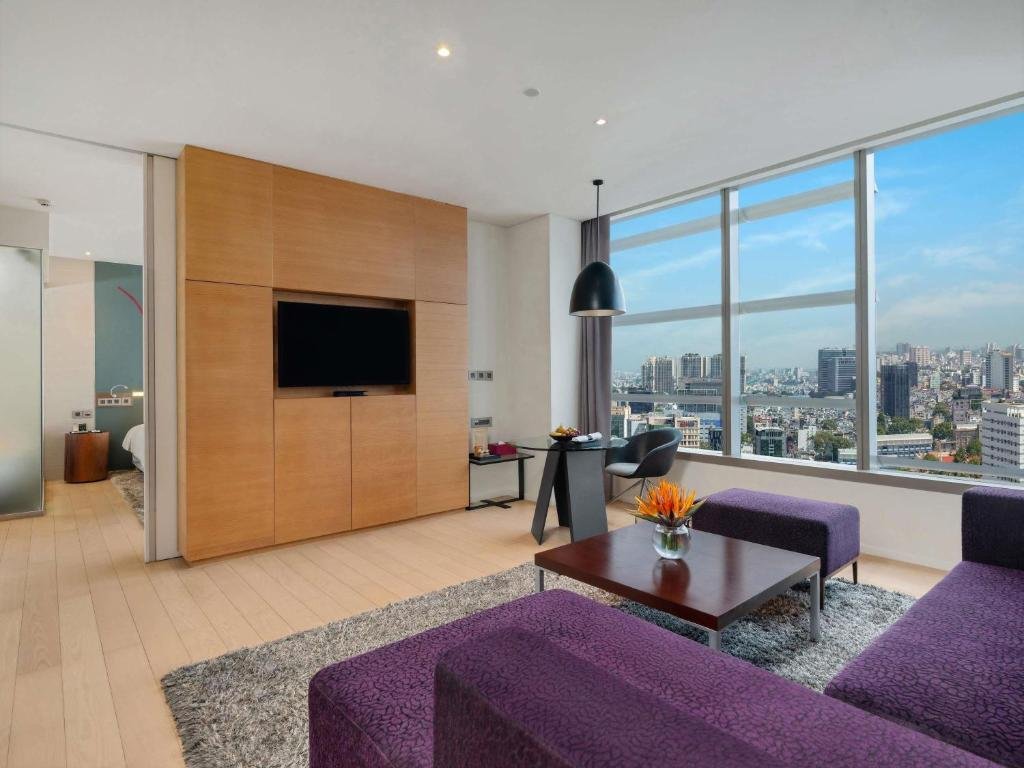 Purple sofas and ottoman around a dark coffee table with a bird-of-paradise arrangement, an oak TV wall, a glass desk by a wide corner window, and the bedroom through a doorway on the left
