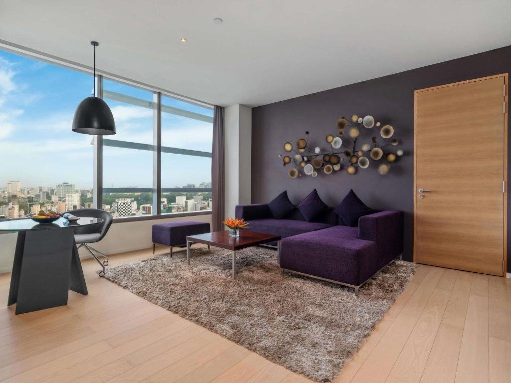 A purple chaise sofa and footstool on a shaggy rug under a disc sculpture, a round glass desk with a fruit bowl by the window, and a light oak floor
