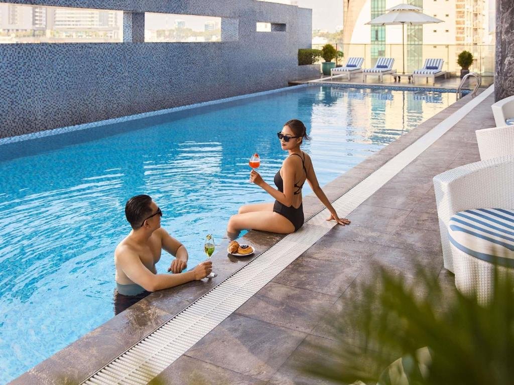 Two guests at the edge of the outdoor pool with cocktails and pastries, striped loungers and an umbrella on the terrace behind