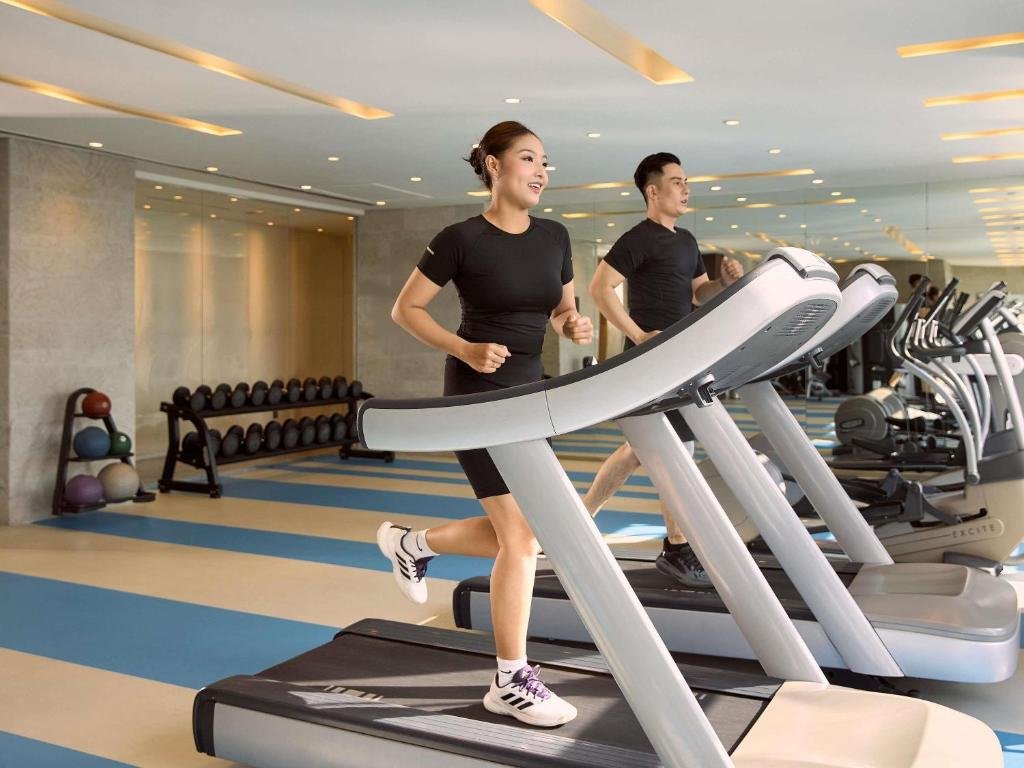 Two guests running on treadmills in a bright gym with a rack of dumbbells and medicine balls against a mirrored wall