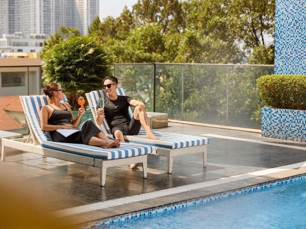 Two guests with cocktails on blue-and-white striped loungers beside the pool, a glass balustrade and trees behind