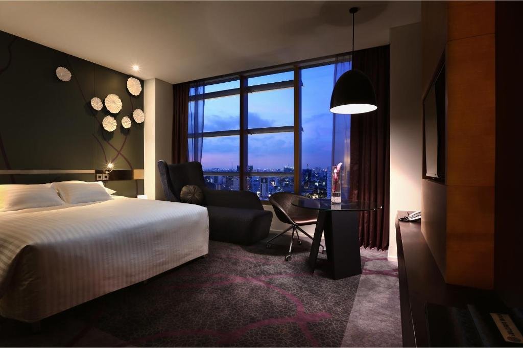 The same room at dusk: the bed, a dark armchair and the glass desk under a black pendant lamp, with the lit city skyline through the full-height window