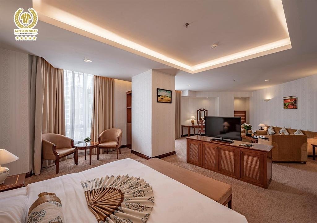 The Suite seen from the foot of the bed, a paper fan laid on the white sheets, with a TV cabinet, two armchairs at the tall curtained window and the sofa area beyond