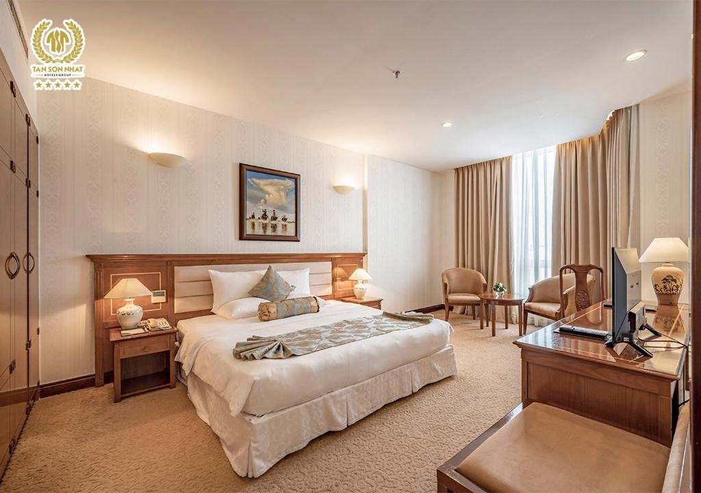 The Suite Deluxe bedroom seen from beside the wardrobe: the bed on the left, a desk with a TV and lamp on the right, and armchairs at the window beyond