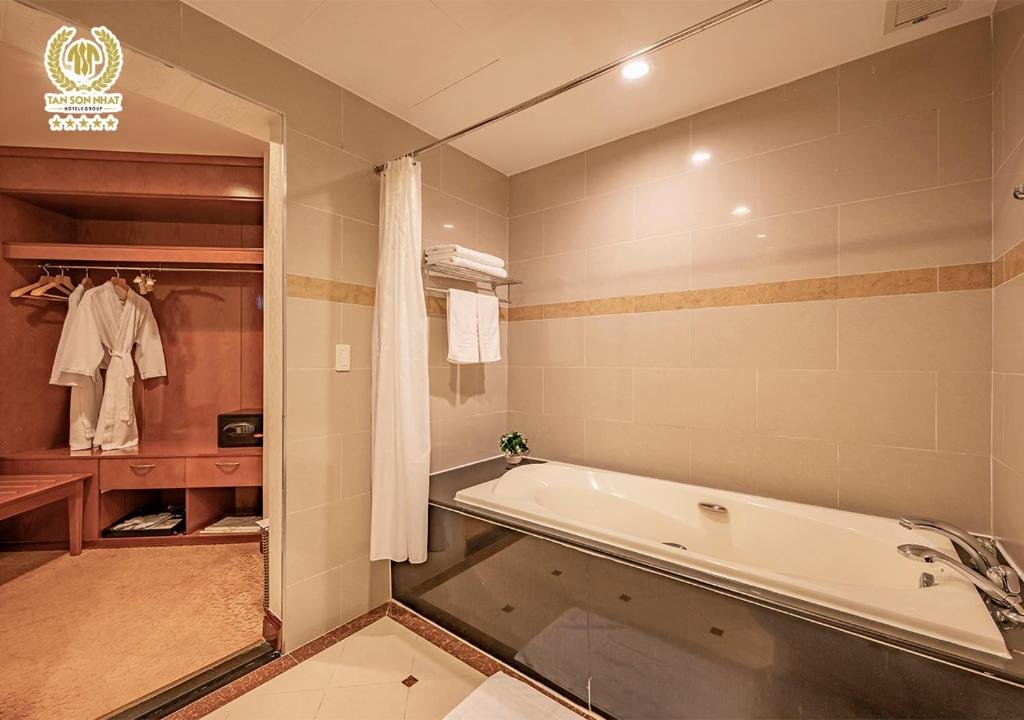 The Suite Deluxe bathroom with a bathtub set in dark stone and a shower curtain, and through the doorway a wooden dressing room with two bathrobes hanging and a safe