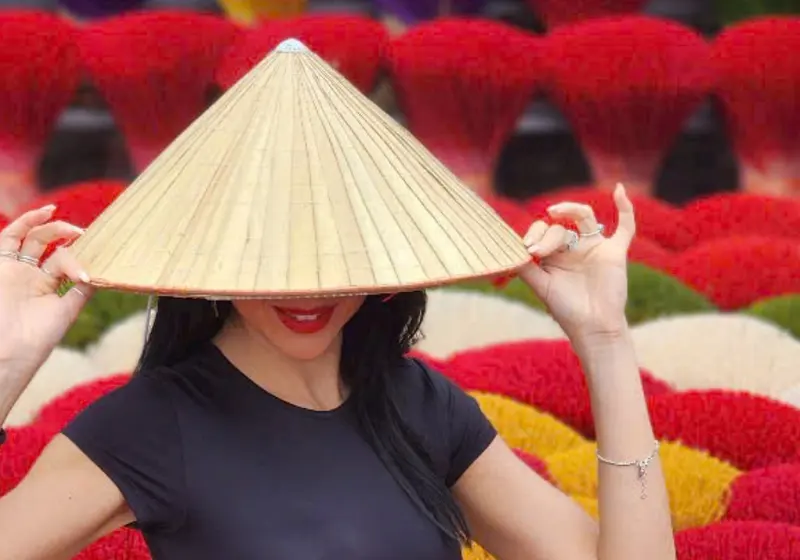 A visitor holding up a conical hat with colourful incense fans behind
