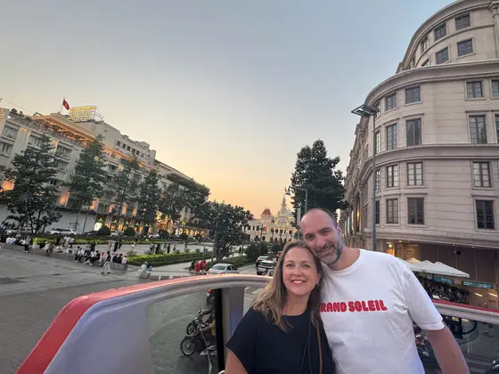 Two guests on the open top deck of the double-decker bus at dusk, the People&rsquo;s Committee building lit behind them