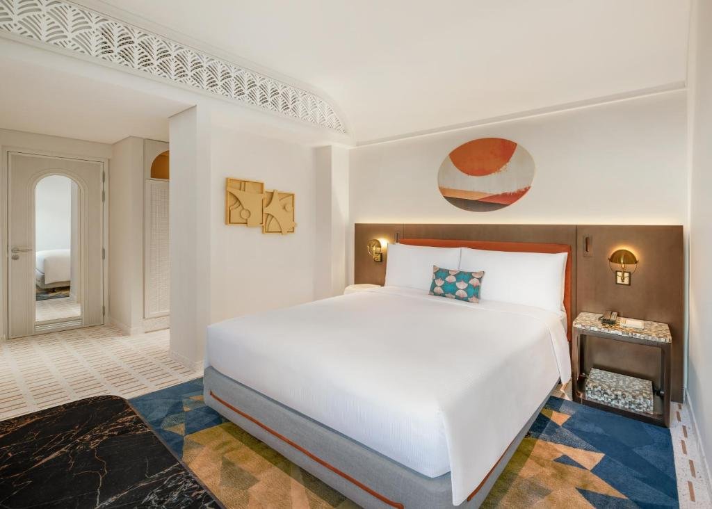 Classico room at La Festa: a king bed in white linen below a carved fretwork arch, an orange and blue abstract canvas above the headboard, and a patterned carpet underfoot