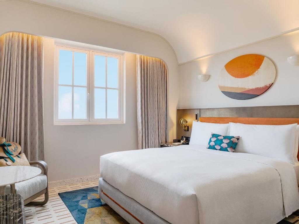 Classico Ocean View at La Festa: a king bed in white linen under a curved vaulted ceiling, a white-framed casement window open to the sky beside it, and a woven sun hat left on the armchair