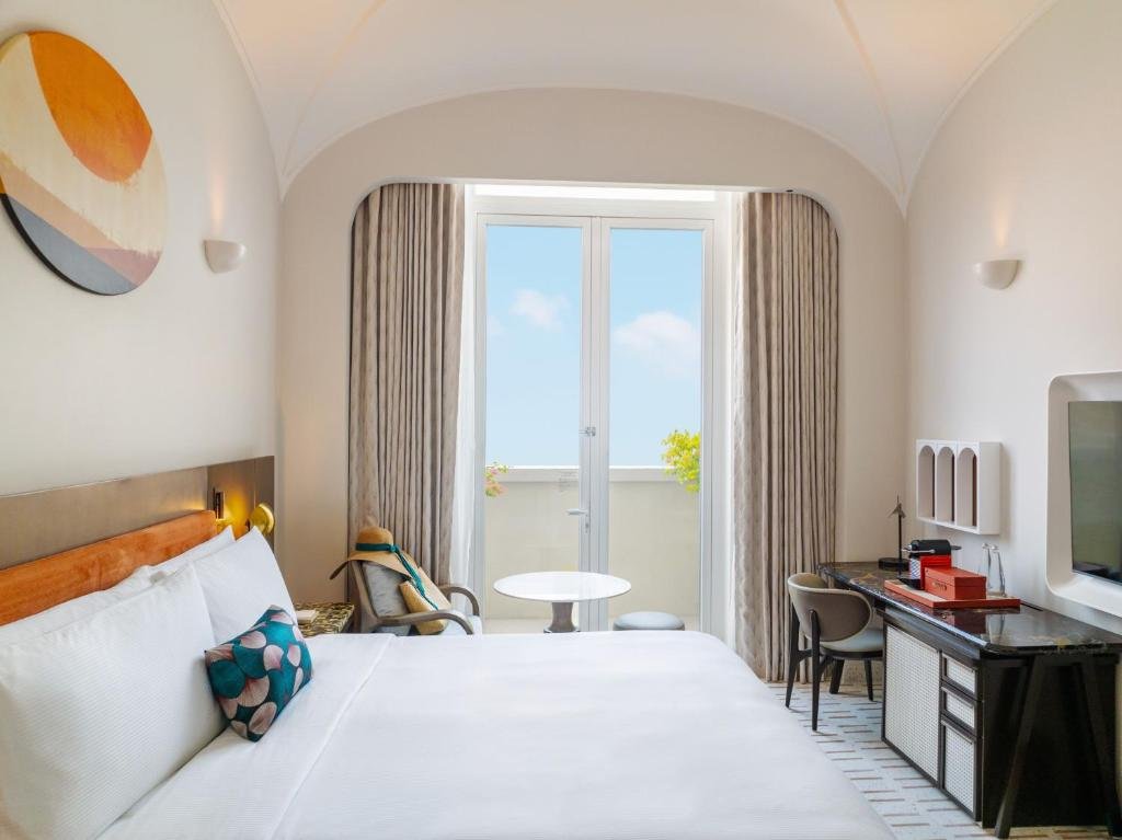 King Room with Balcony at La Festa: a king bed under a vaulted ceiling facing French doors onto a balcony set with a small round table, a desk and television along the right-hand wall, and a sun hat left on the armchair