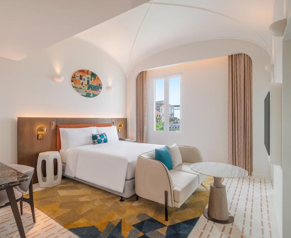 King Room with Balcony and Sea View at La Festa: a king bed under a vaulted ceiling with a round harbour-village canvas above it, a casement window onto the terracotta roofs and palms of Sunset Town, and an armchair with a marble side table