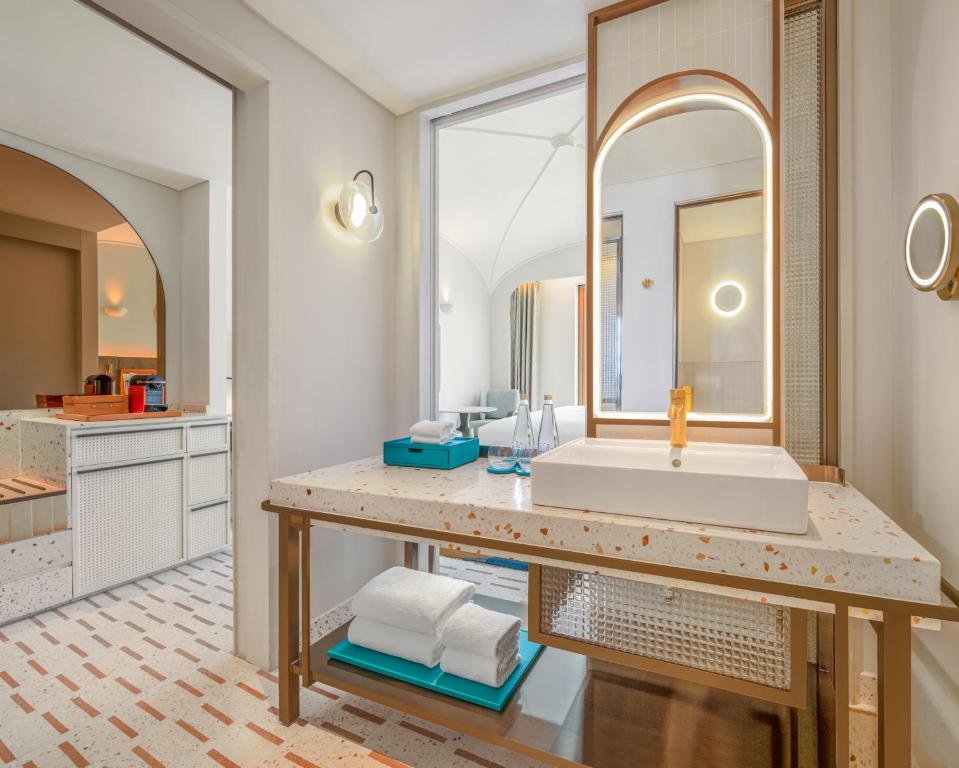 The bathroom: a terrazzo counter on a brass frame with a square basin and gold tap, a backlit arched mirror above and folded towels on the shelf below, with the bedroom visible through the doorway