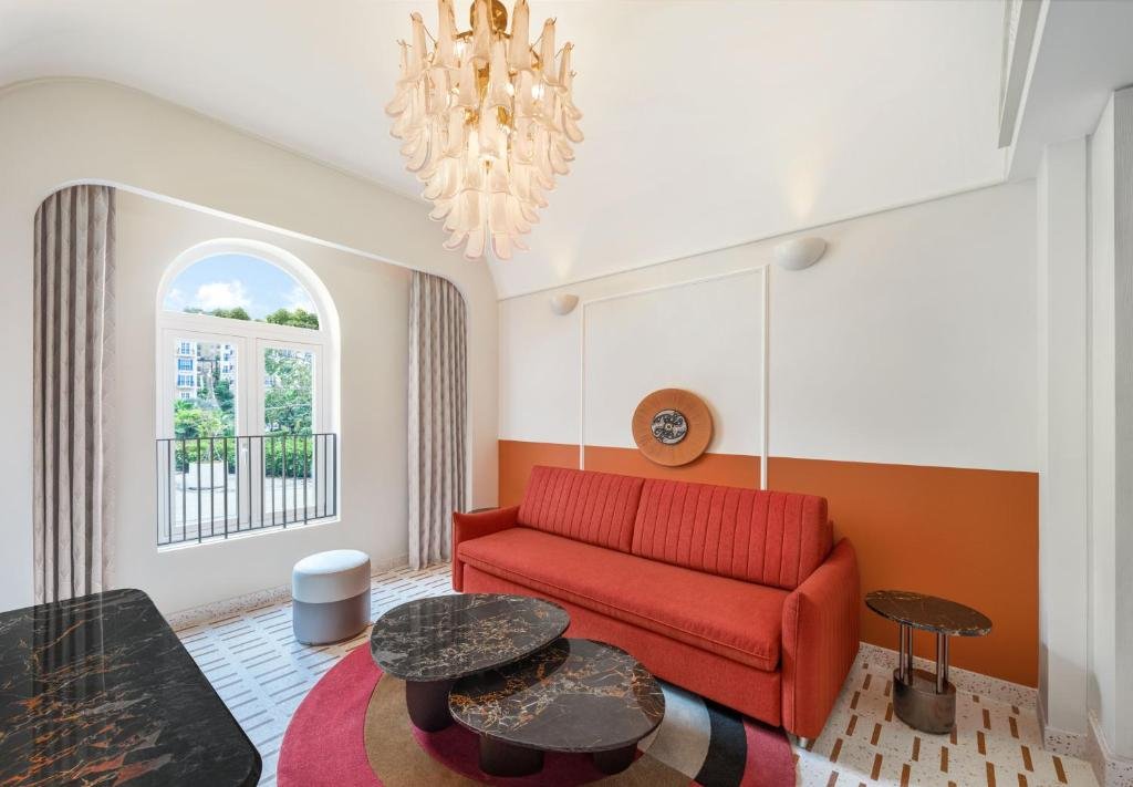 The Deluxe King Room sitting area: a coral sofa below an orange dado, a glass petal chandelier overhead, marble-topped nesting tables on a round rug, and an arched window onto a Juliet balcony