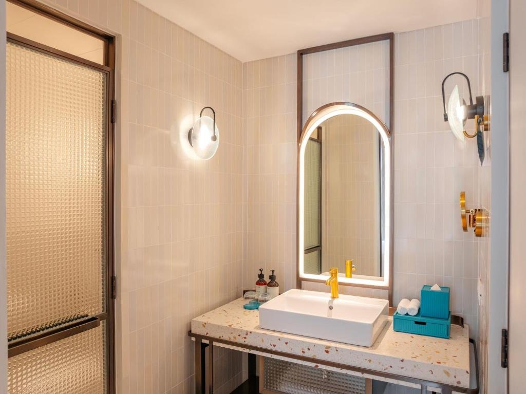 The Deluxe King Room bathroom: a square basin and gold tap on a terrazzo counter under a backlit arched mirror, with a walk-in shower behind a textured glass door