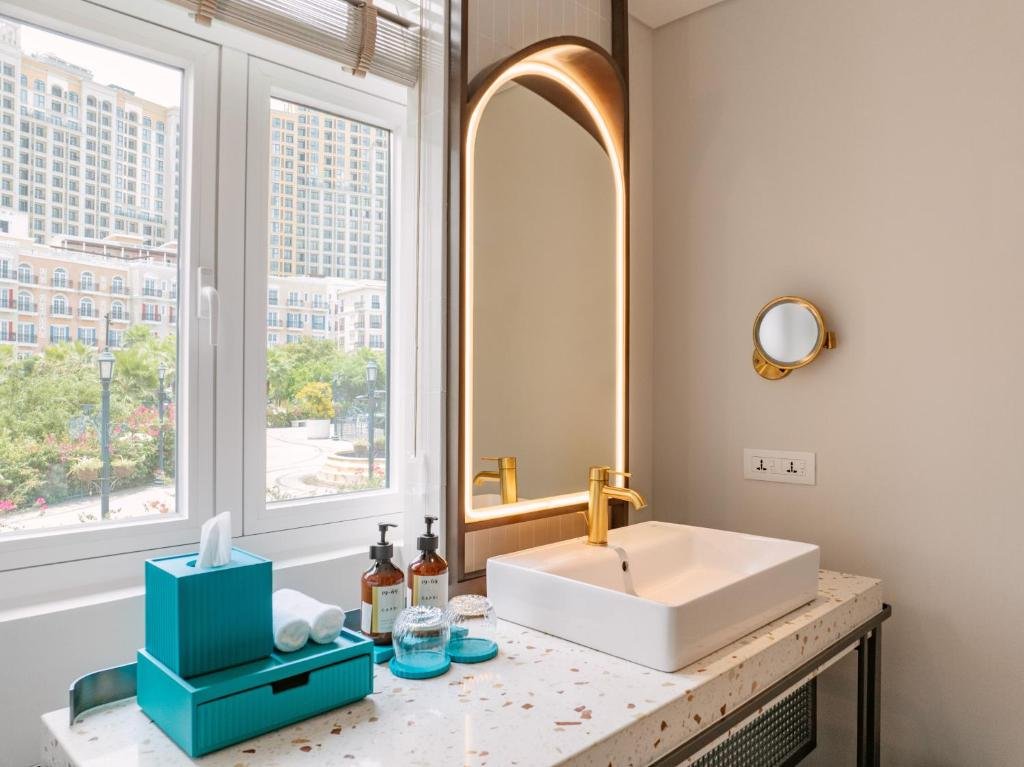 The Classico bathroom vanity: a square basin on a terrazzo counter with a gold tap and a backlit arched mirror, beside a window looking onto the buildings and gardens of Sunset Town