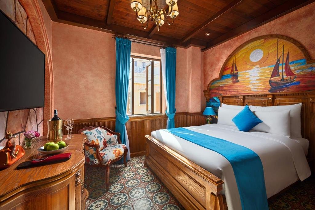 Caeli Amoris at Venice Hotel: a double bed under an arched mural of sailing boats at sunset, a wood-beamed ceiling with a brass chandelier, a window hung with blue curtains, a floral armchair on patterned floor tiles, and a TV above a carved console