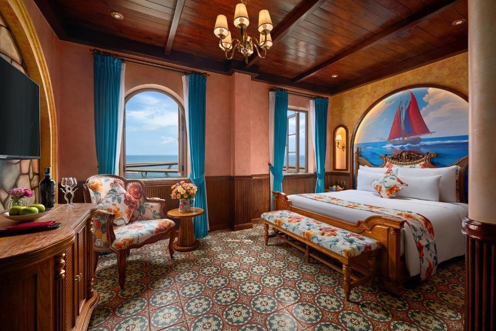 Mare Amoris at Venice Hotel: a large double bed under an arched mural of a red-sailed ship, two arched windows hung with blue curtains, one open onto the sea, and a floral armchair and a bench at the foot of the bed on patterned floor tiles