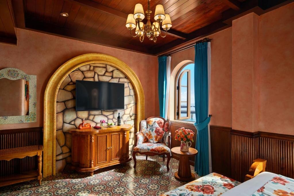 A TV set into a stone-clad arch above a carved console, a mosaic-framed mirror, and a floral armchair beside an arched window with the sea beyond