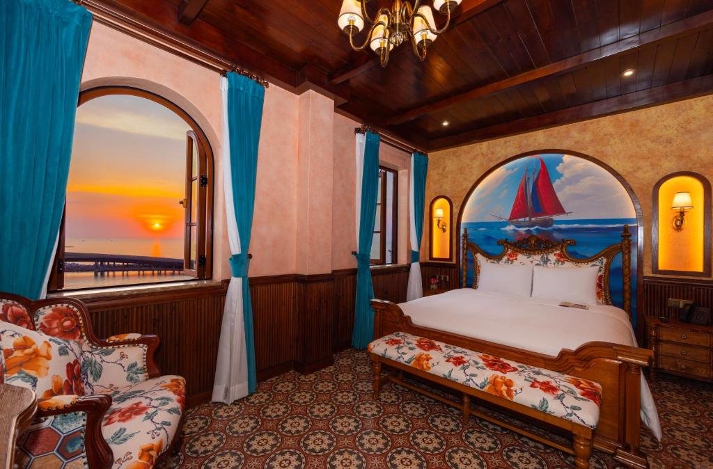 The sun setting over the sea through an arched window, a footbridge running out over the water, with the bed under its red-sailed ship mural and a floral bench on the right