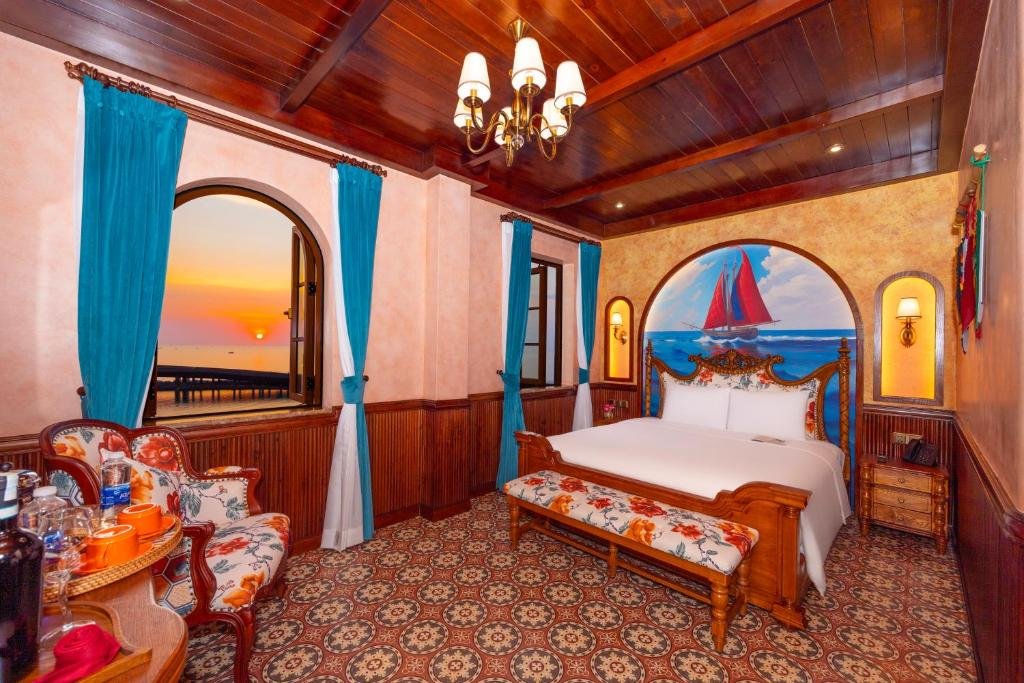 The room at sunset from beside the tea tray: floral armchairs, the arched window glowing with the sun going down over the sea, and the bed under the ship mural