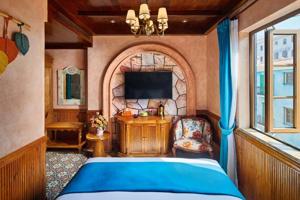 The room seen from the bed: a TV set into a stone-clad arch above a carved console, a floral armchair, and a bay window opening onto the pastel buildings of Sunset Town