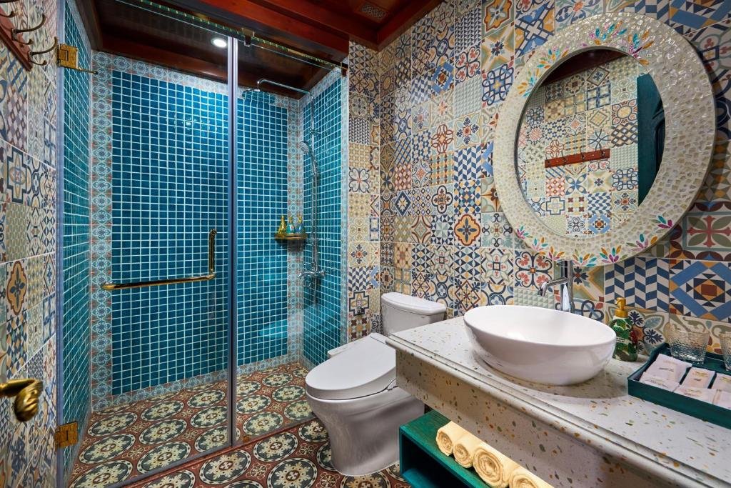 The bathroom: a walk-in shower lined with blue mosaic tile behind glass, walls of patterned encaustic tile, a round mosaic-framed mirror over a vessel basin on terrazzo, and a toilet