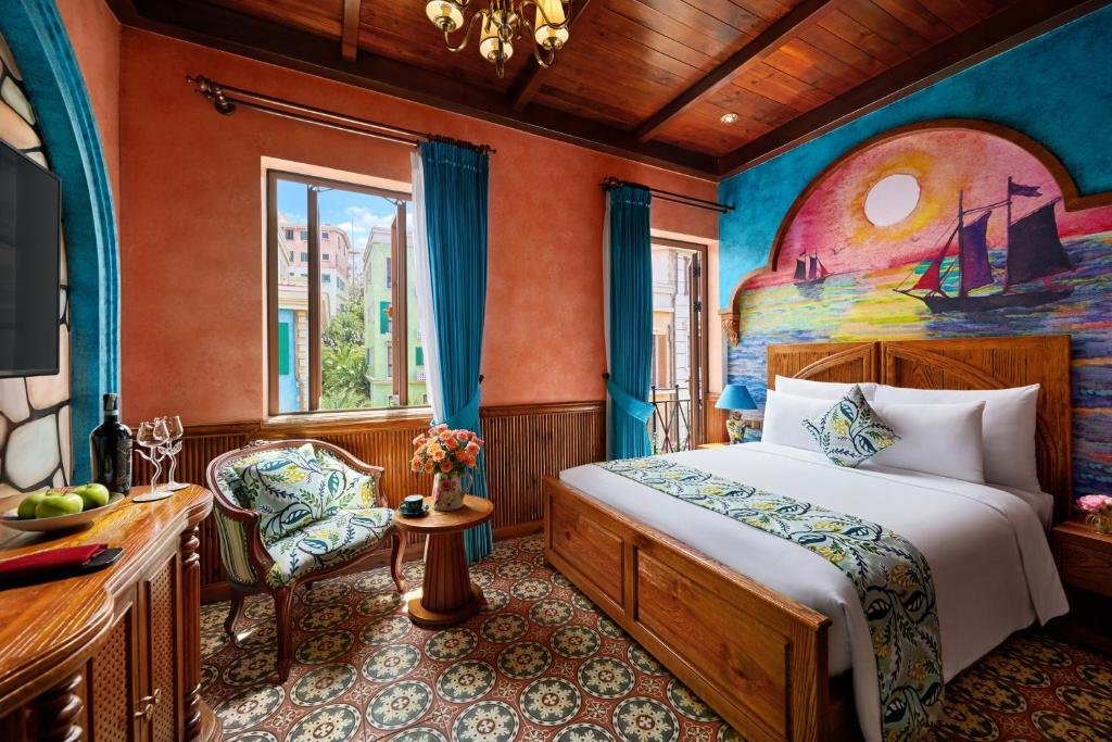 Lumen Amoris at Venice Hotel: a double bed under a sunset mural of sailing ships on a blue wall, a window and a glazed balcony door hung with blue curtains, and a leaf-print armchair with a round table on patterned floor tiles