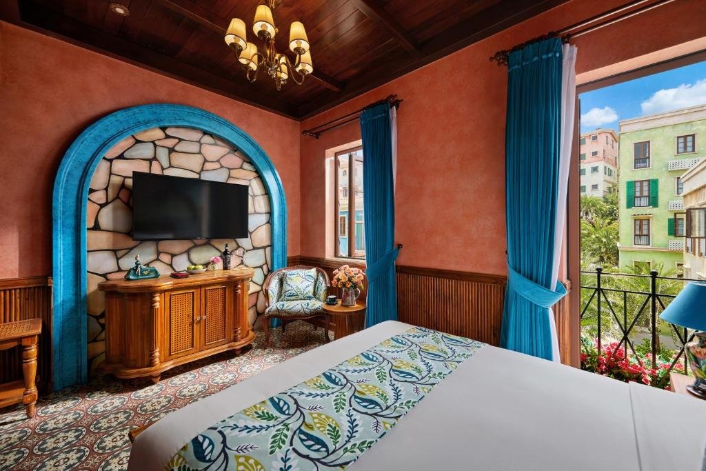 The room from the bed: a TV in a stone-clad arch trimmed in blue above a carved console, and the balcony door standing open onto a wrought-iron railing with flowers and the pastel buildings of Sunset Town