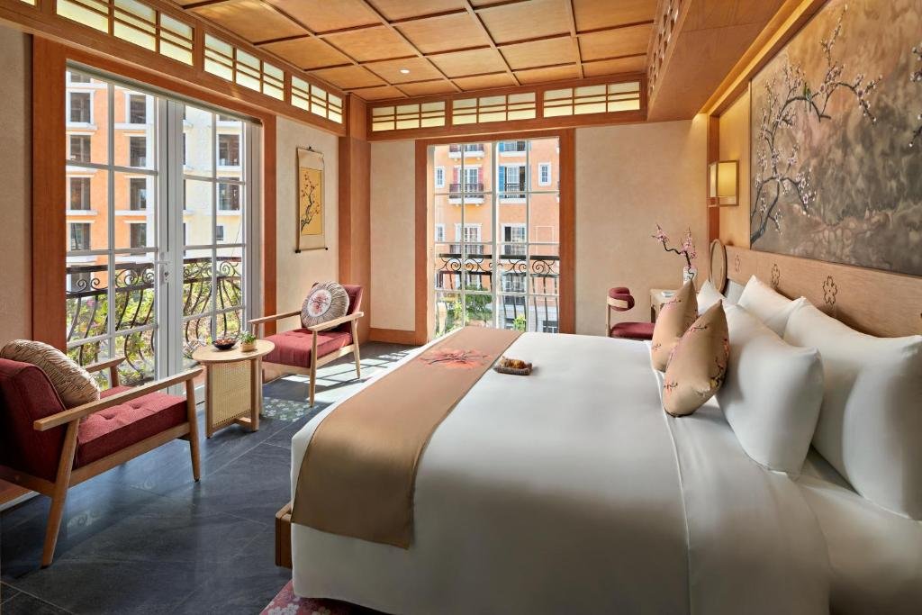 Yukizakura at Sakurakyo: a king bed under a painted blossom panel and a coffered wood ceiling, with two sets of French doors onto wrought-iron balconies looking over the pastel buildings of Sunset Town, and a pair of armchairs by the windows