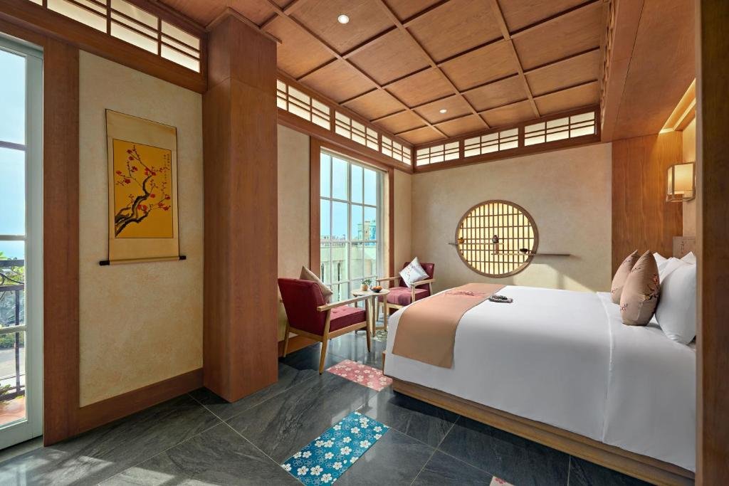 Red armchairs and a round table by a tall window, the round lattice window beside the bed, a yellow blossom scroll, and a glazed door onto the balcony at the left