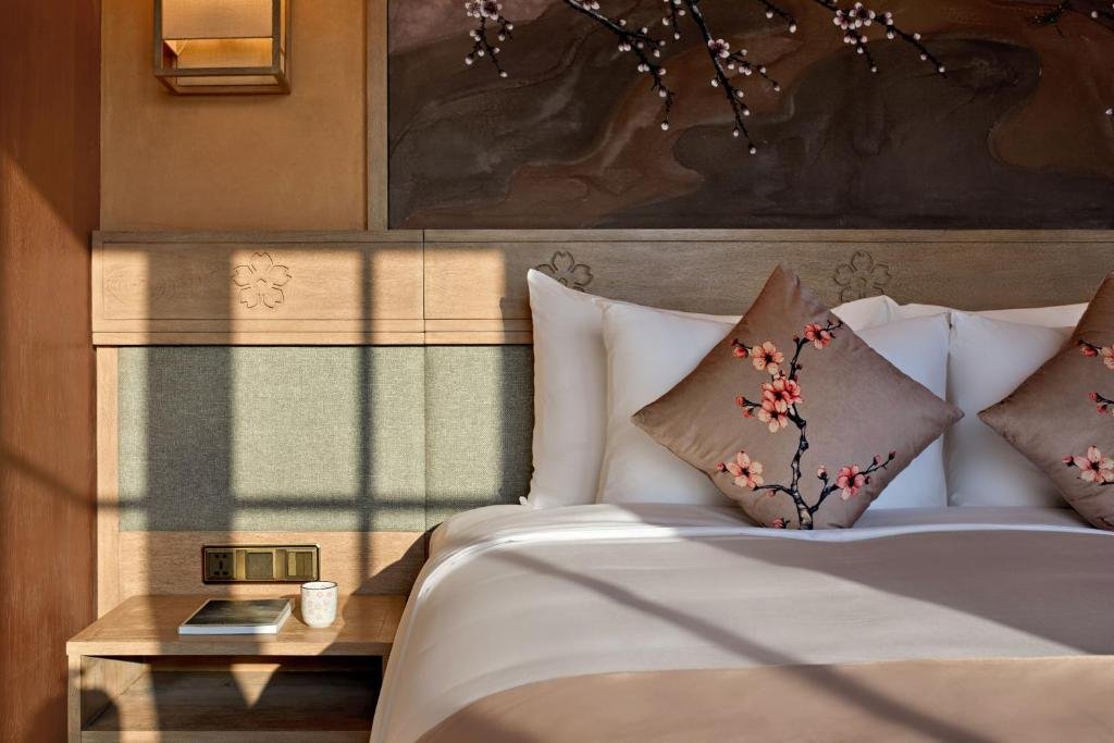 Close view of the bed: embroidered blossom cushions, a headboard carved with cherry-blossom motifs, a bedside table with a book and a cup, and sunlight falling in window-pane shapes