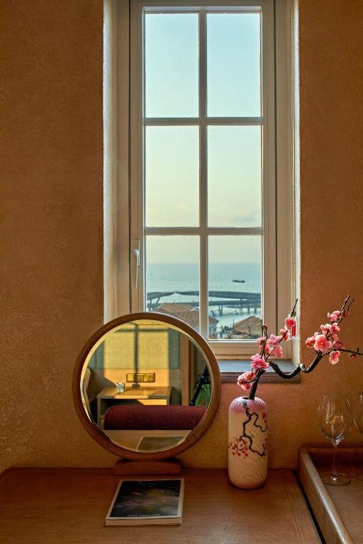 A tall window looking out over the sea to a long walkway on the water, above a dressing table with a round mirror and a vase of blossom