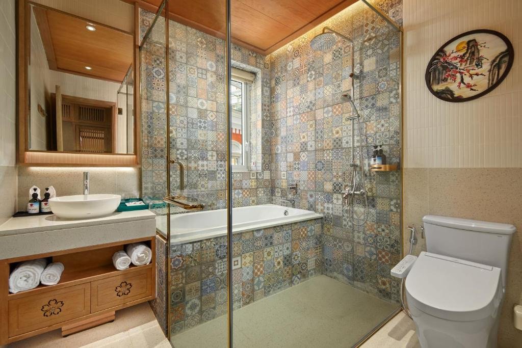 A bathtub set inside a glass-walled shower lined with patterned encaustic tile, a vessel basin on a wooden vanity carved with blossoms, and a toilet