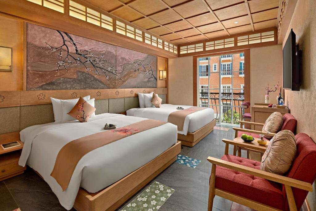 Two beds, a double and a large double, side by side beneath a long blossom mural, with red armchairs and a round table by a window onto the pastel buildings of Sunset Town, and a TV on the wall