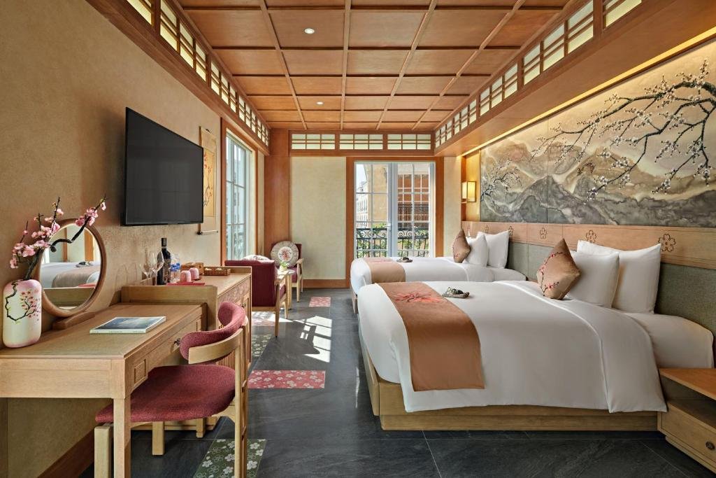 Two beds, a double and a large double, beneath a long blossom mural, with a dressing table and wall TV opposite and a glazed door onto a wrought-iron balcony at the far end