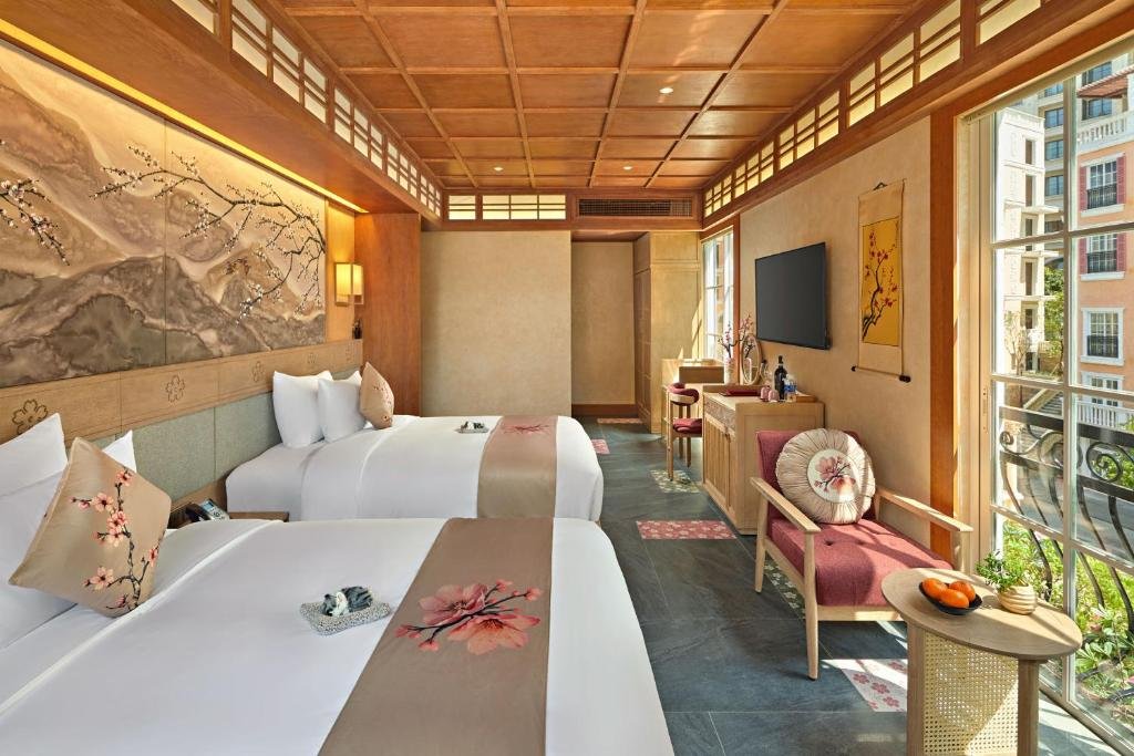 The room from the window end: both beds along the mural wall, a wall TV and minibar console at the far side, and an armchair with a round table beside floor-to-ceiling windows onto the balcony