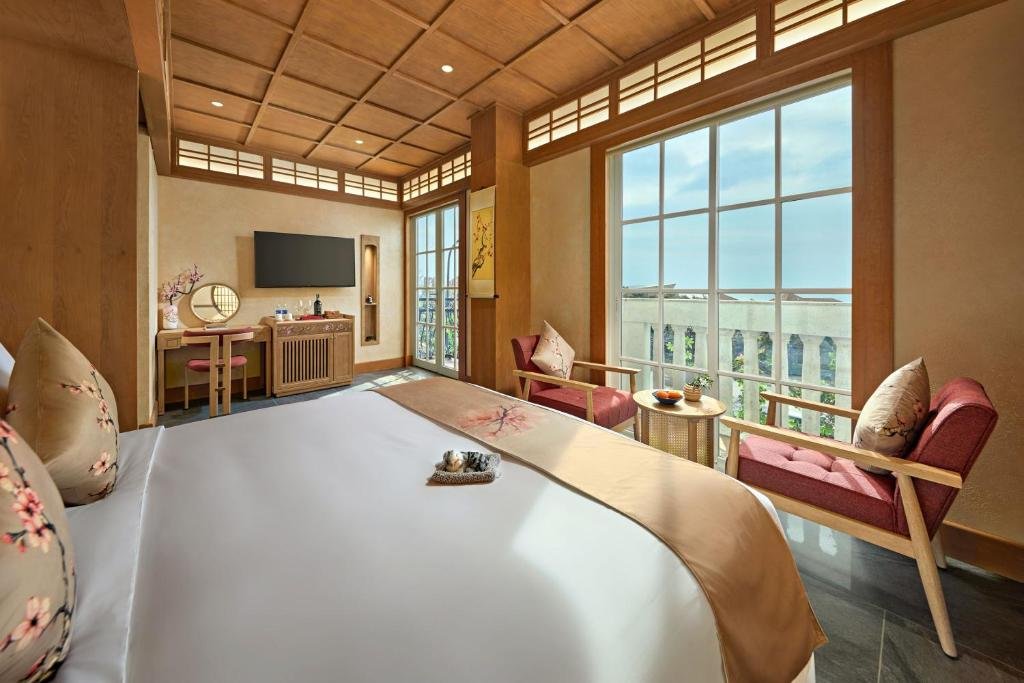 A king bed facing a wall of windows onto a balustraded terrace, two armchairs and a round table by the glass, and a dressing table with a round mirror beneath a wall-mounted TV