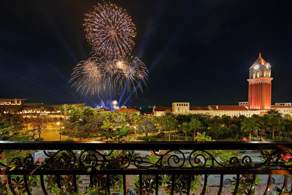 Fireworks bursting over Sunset Town at night, seen from a wrought-iron balcony, with the lit clock tower on the right and the palms of the square below
