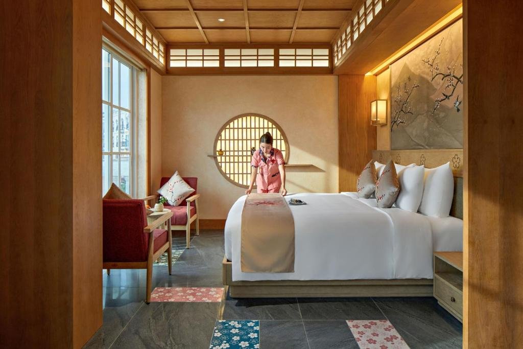 A housekeeper smoothing the bed runner in a room with a round lattice window, a blossom mural above the bed and red armchairs by the tall window