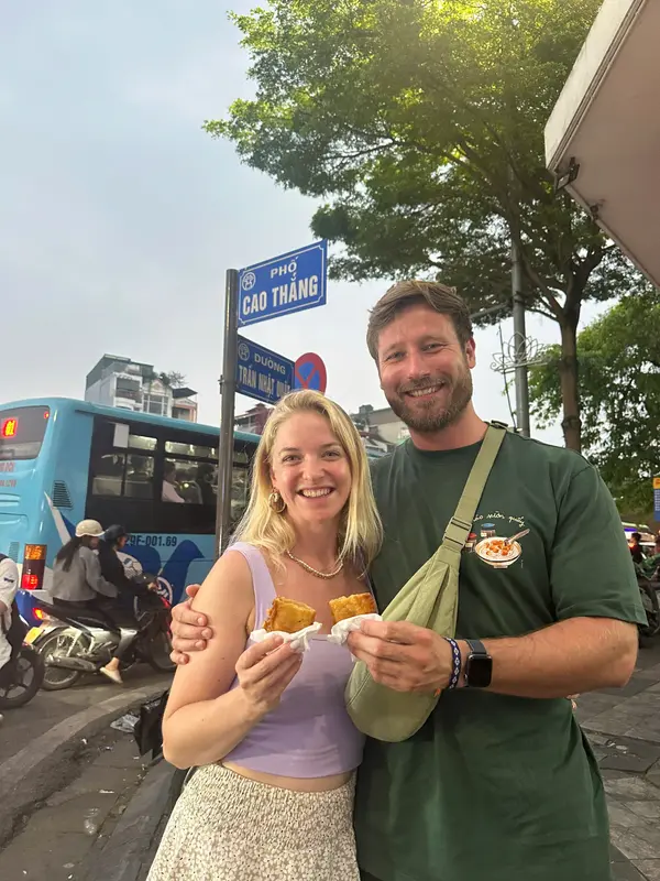 Two guests holding fried pastries at a Hanoi street corner under the Pho Cao Thang sign