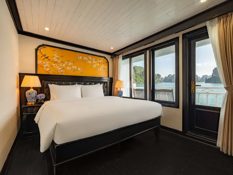 Deluxe Private Balcony cabin on deck 2: double bed, gold blossom headboard panel and a glass door opening onto a private balcony over the bay