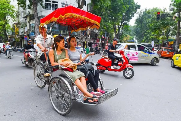 Two guests riding side by side in a cyclo through Hanoi traffic, their driver pedalling behind