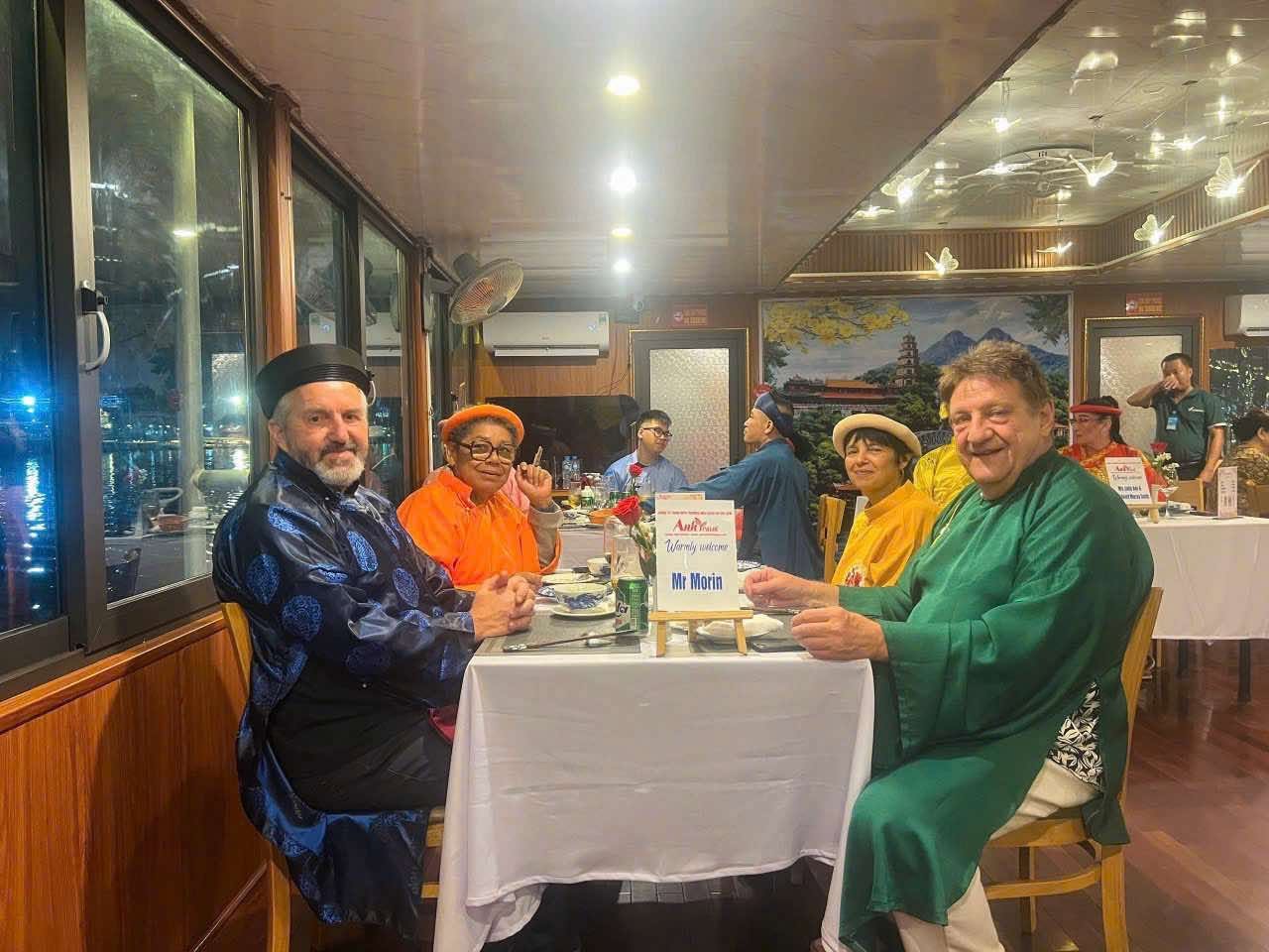 Four guests in Huế court robes at their table, the river lights behind them